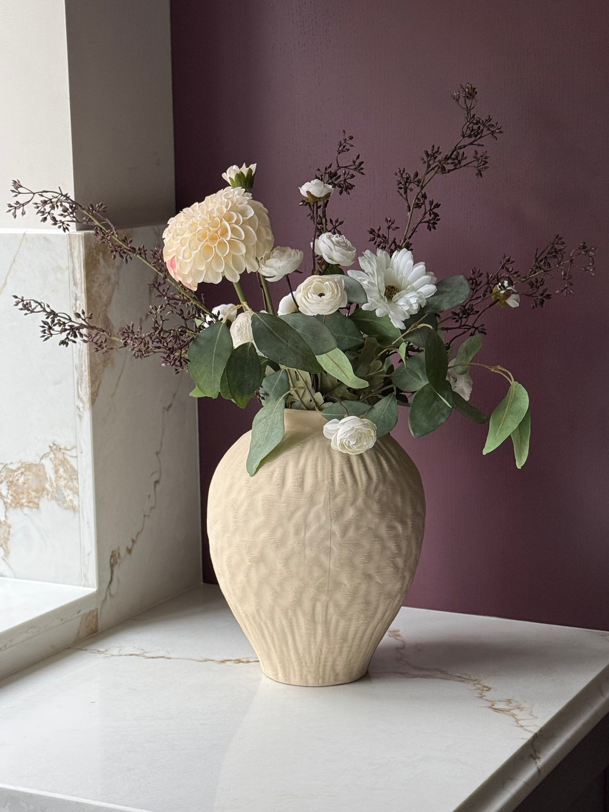 A textured cream ceramic vase filled with white and cream flowers and green foliage, displayed on a white marble ledge against a dark purple wall.