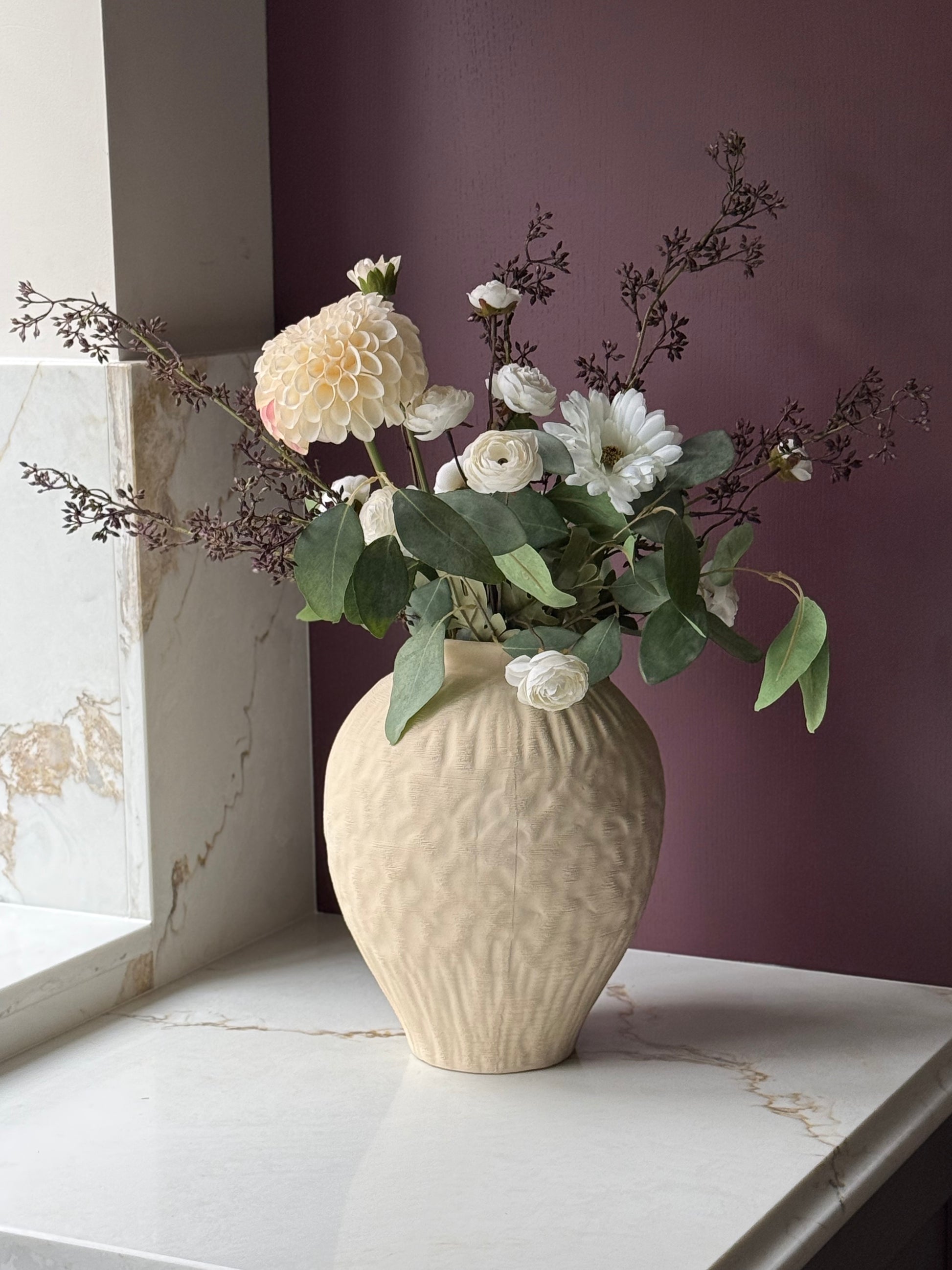 A textured cream ceramic vase filled with white and cream flowers and green foliage, displayed on a white marble ledge against a dark purple wall.