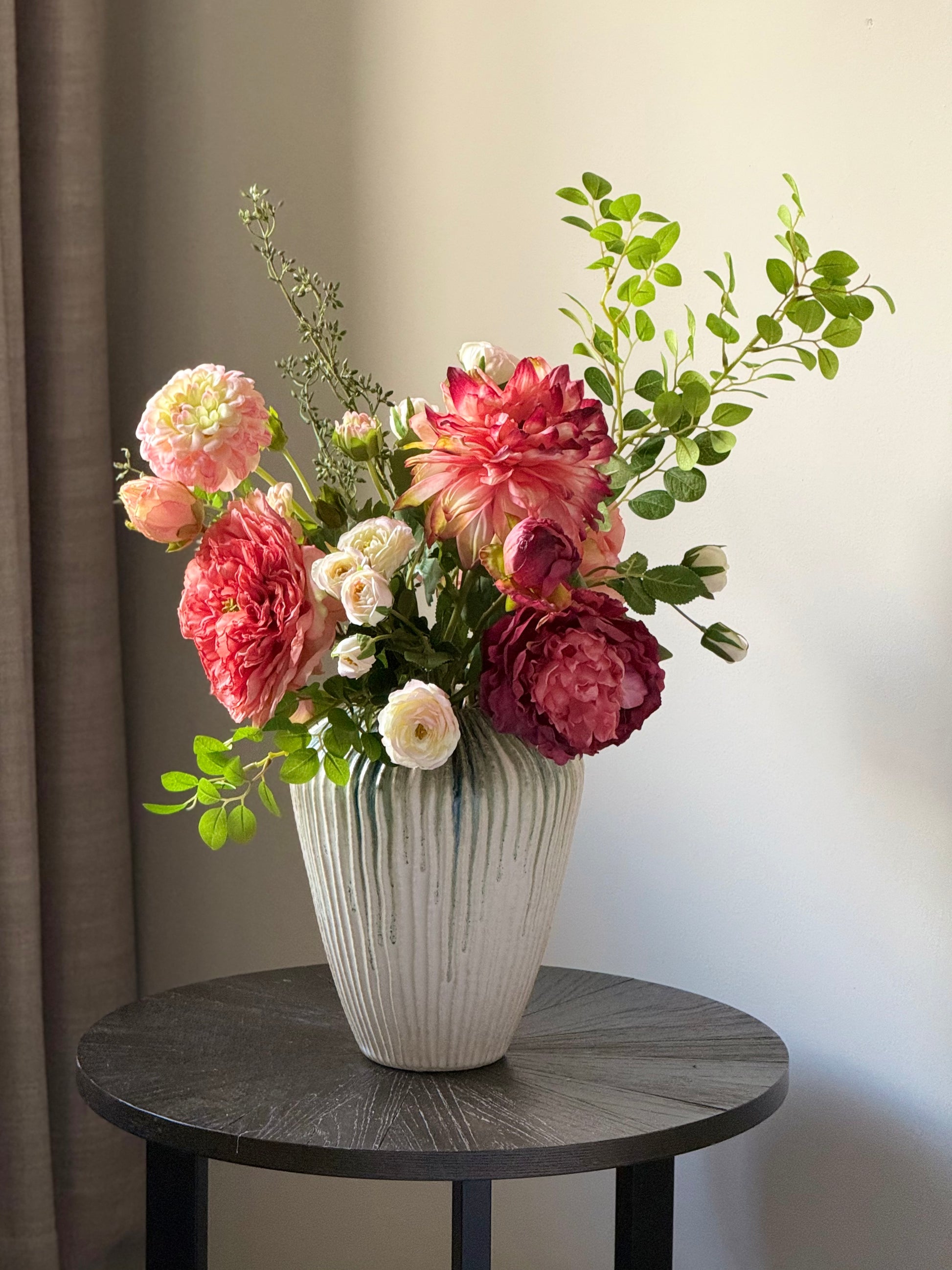 luxury artificial red and coral Floral arrangement in a white vase on a round wooden table against a neutral wall.