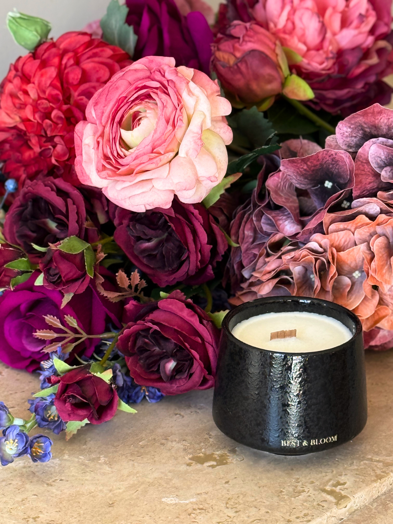 Black Best & Bloom candle with a wooden wick on a stone surface, surrounded by vibrant artificial flowers.