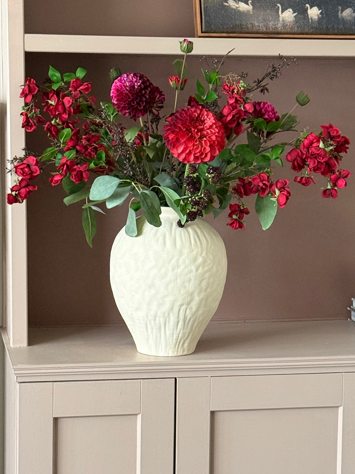 A textured off-white ceramic vase filled with a vibrant floral arrangement of red and magenta dahlias and green foliage, displayed on a light beige cabinet against a warm brown wall.