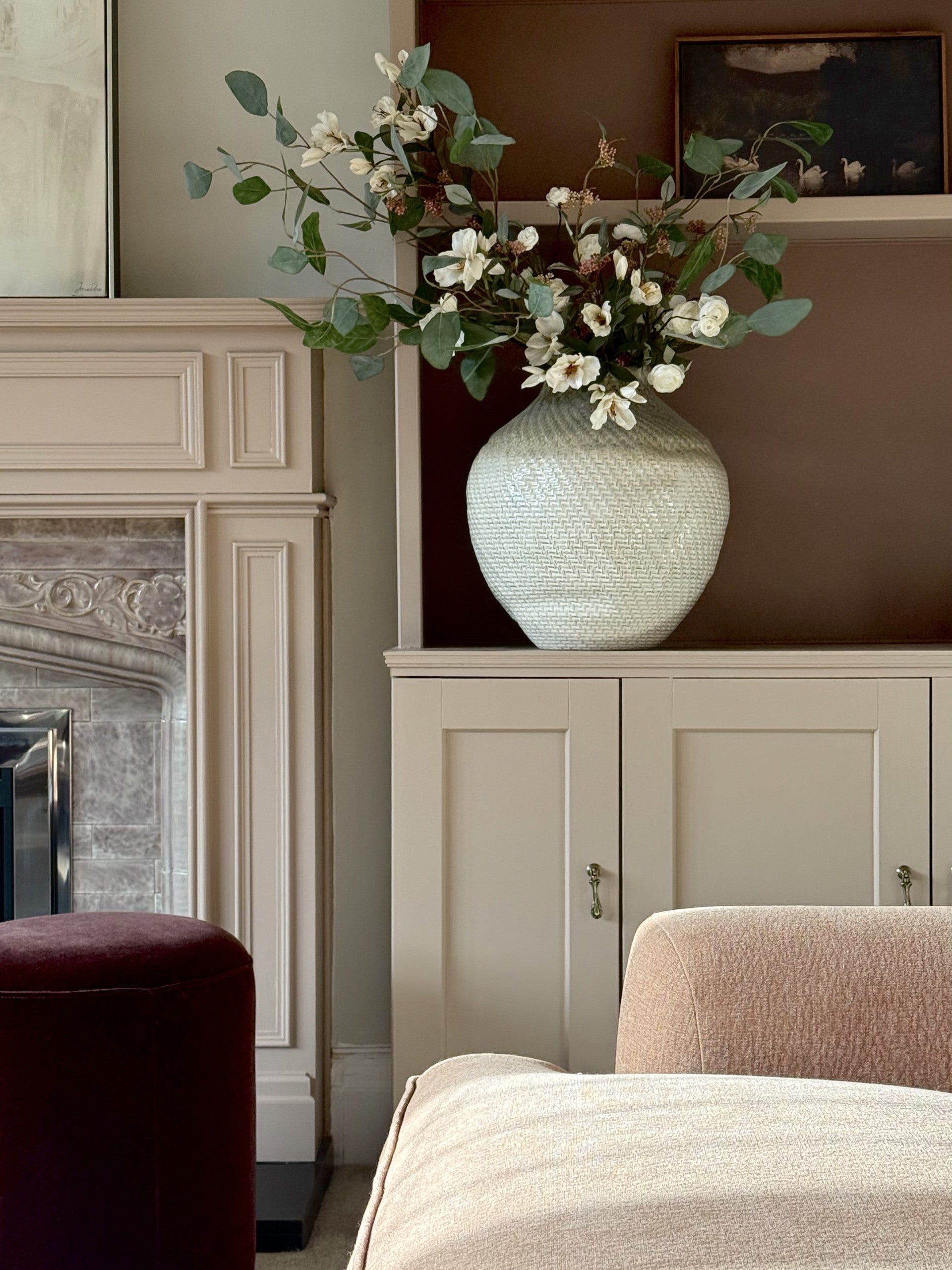 A large textured cream ceramic vase filled with white flowers and green eucalyptus stems, displayed on a cream cabinet in a styled living room next to a fireplace.