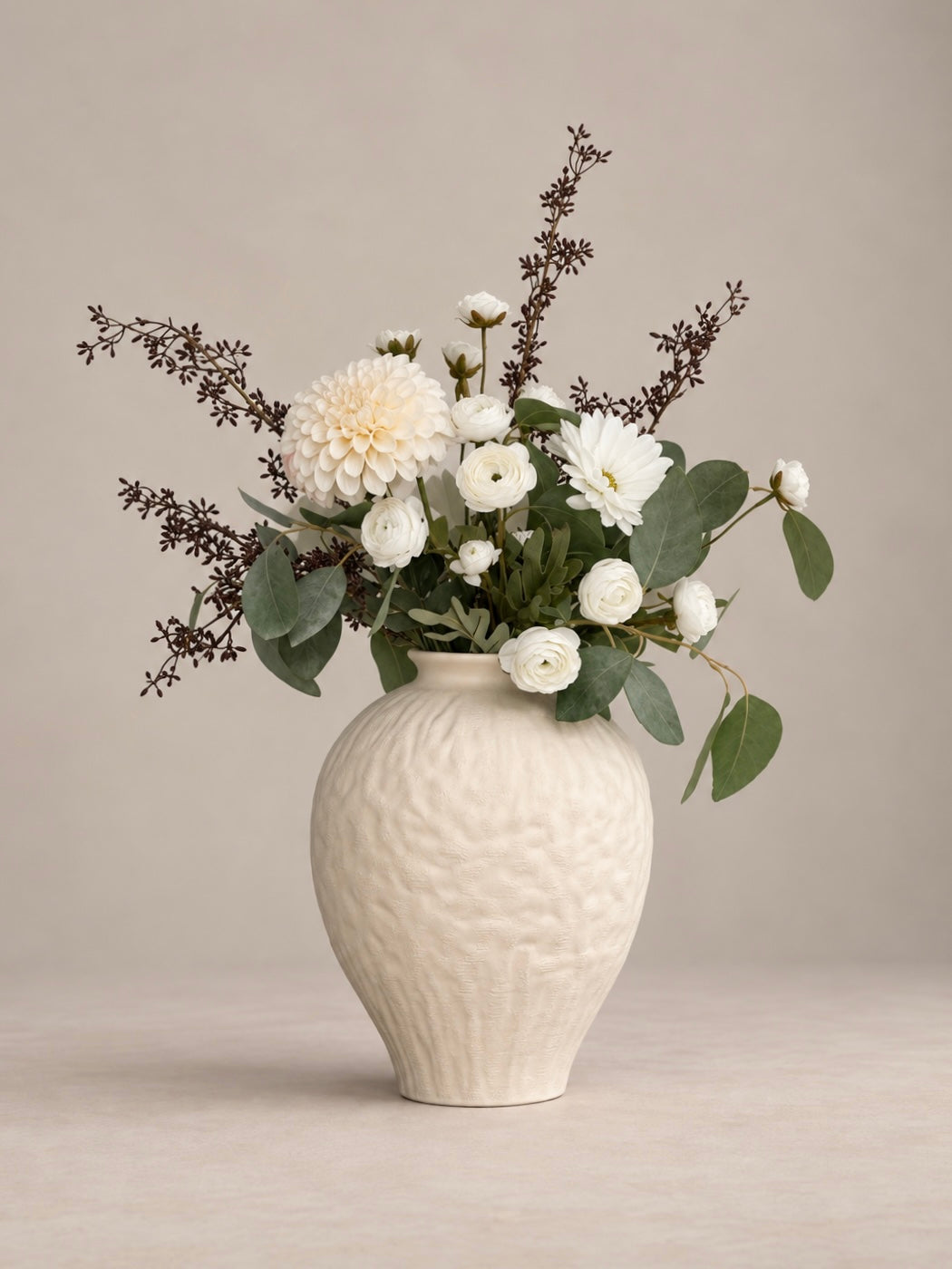 A textured cream ceramic vase filled with a floral arrangement of white dahlias, white ranunculus, green eucalyptus leaves, and dark brown filler stems, set against a neutral beige background.
