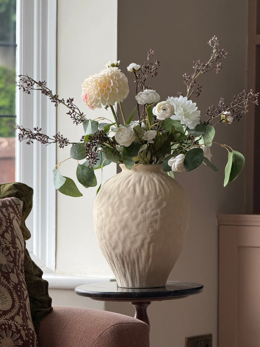 A cream-colored textured ceramic vase filled with a floral arrangement of white and cream dahlias, roses, and green foliage, displayed on a small round table in a sunlit room.