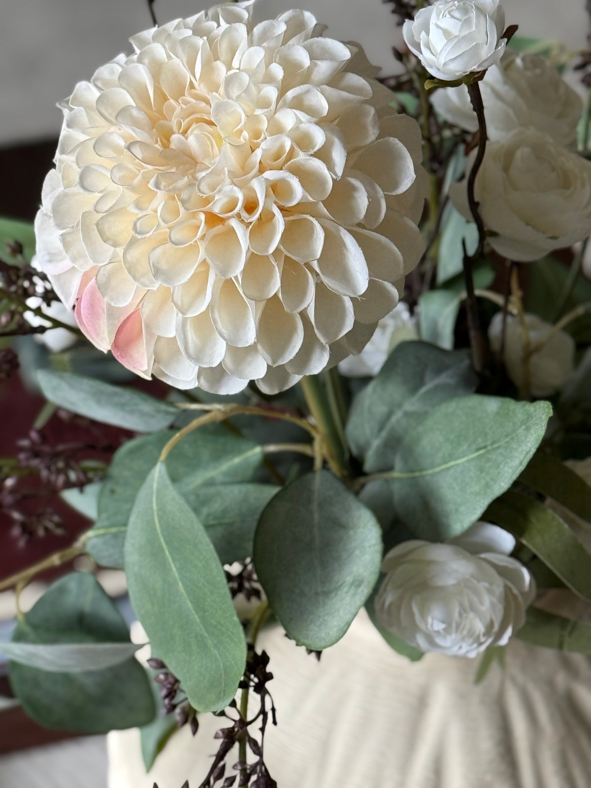 A close-up of a floral arrangement featuring a large cream dahlia, white ranunculus, green eucalyptus leaves, and a soft pink accent flower.